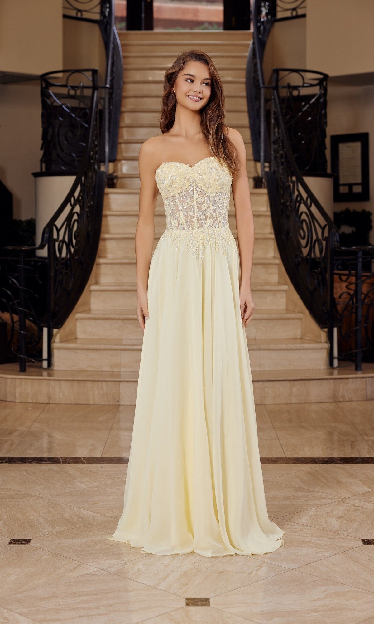 A woman stands on marble flooring by a staircase, wearing the Nox Anabel C1617 strapless sweetheart prom dress in lemon with a sheer embroidered bodice featuring floral details and a flowing skirt.